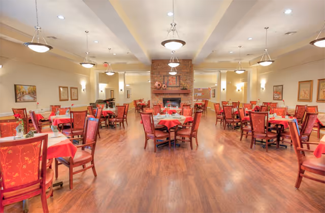 A spacious dining room with multiple round tables covered with red tablecloths and surrounded by red cushioned chairs. The room features wooden flooring, a brick fireplace at the far end, and several framed pictures on the walls. Ceiling lights and wall sconces provide warm lighting.