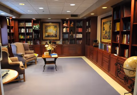 A cozy library or reading room with wooden bookshelves filled with books and decorative items. There are two upholstered armchairs and a small wooden table with a flower arrangement in the center. The room has a carpeted floor and framed artwork on the walls.