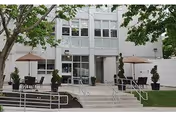 Front entrance of a multi-story building with outdoor patio tables, umbrellas, planters, stairs and a wheelchair ramp.