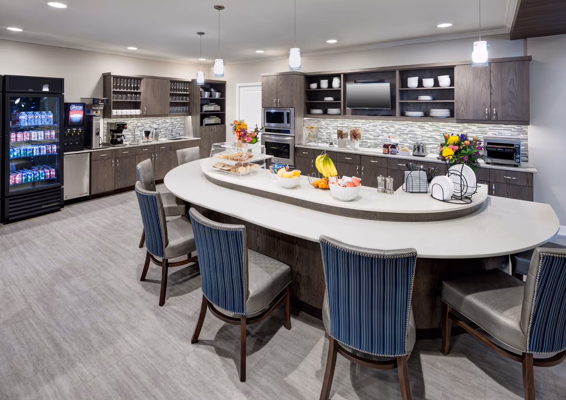 A modern kitchen area with a large curved island countertop surrounded by six upholstered chairs. The kitchen features dark wood cabinets, a tiled backsplash, built-in microwave and oven, a refrigerator stocked with beverages, a coffee machine, and various bowls of fruit and snacks on the island. Pendant lights hang from the ceiling, and there are decorative flowers on the counter.