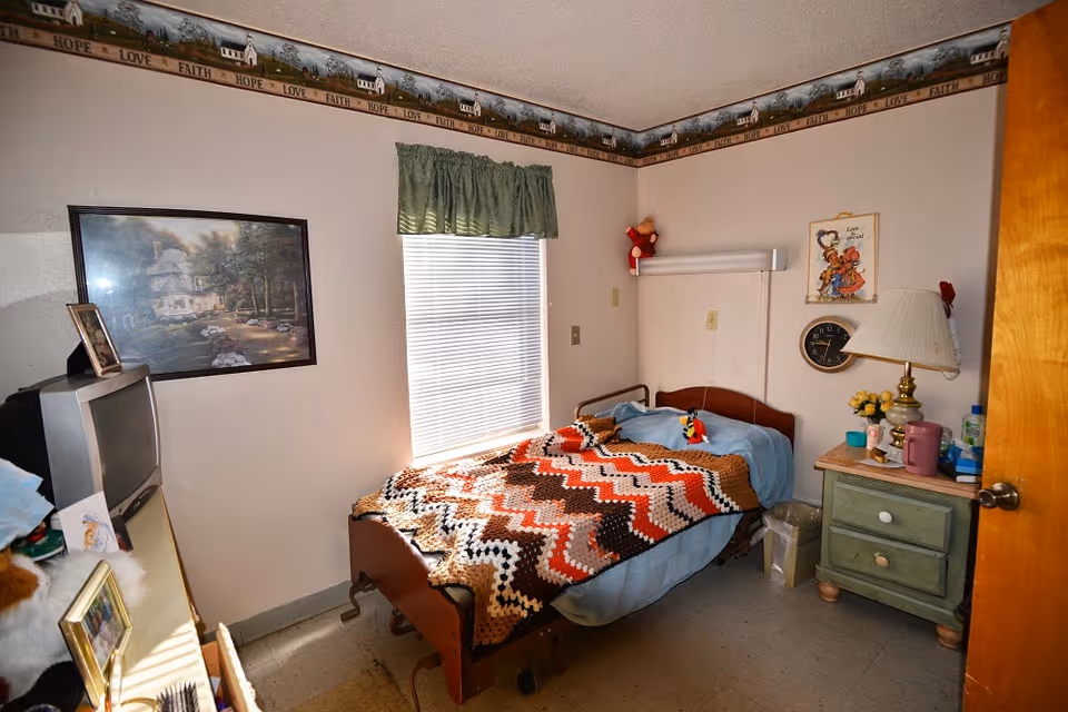 Small nursing home bedroom with a single bed covered by a crocheted blanket, a nightstand, TV and a window.