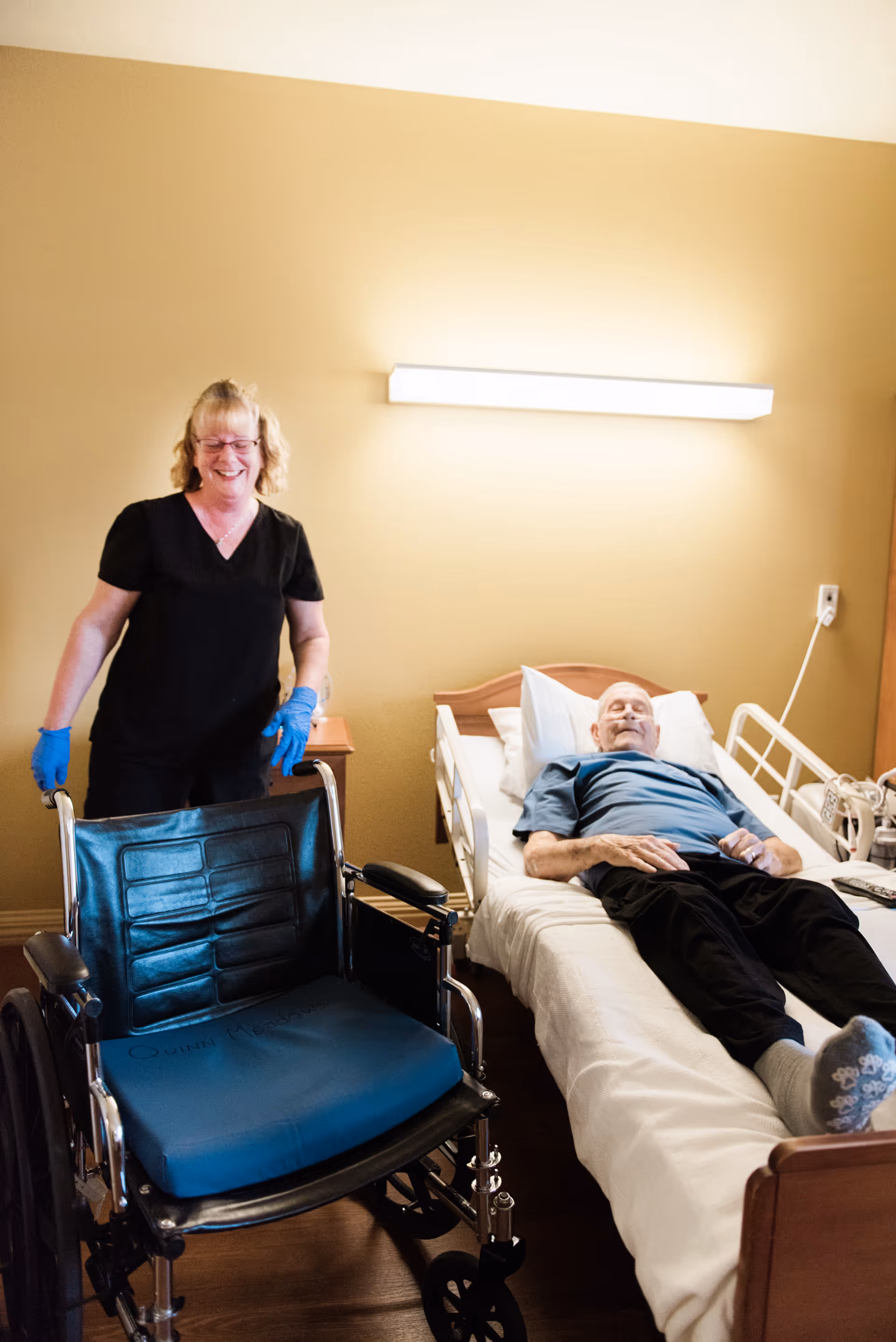 A smiling caregiver stands beside a wheelchair while an elderly man lies in a hospital-style bed in a senior care room.