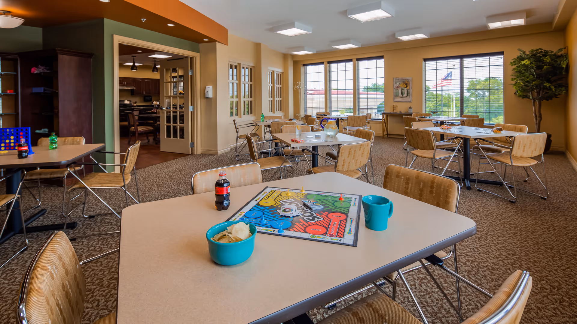 A bright and spacious common room with multiple tables and chairs arranged for group activities. On the nearest table, there is a board game, a bowl of chips, a blue mug, and a bottle of soda. Large windows let in natural light, and there is a potted plant in the corner. The room has carpeted floors and neutral-colored walls with some artwork and decorations.