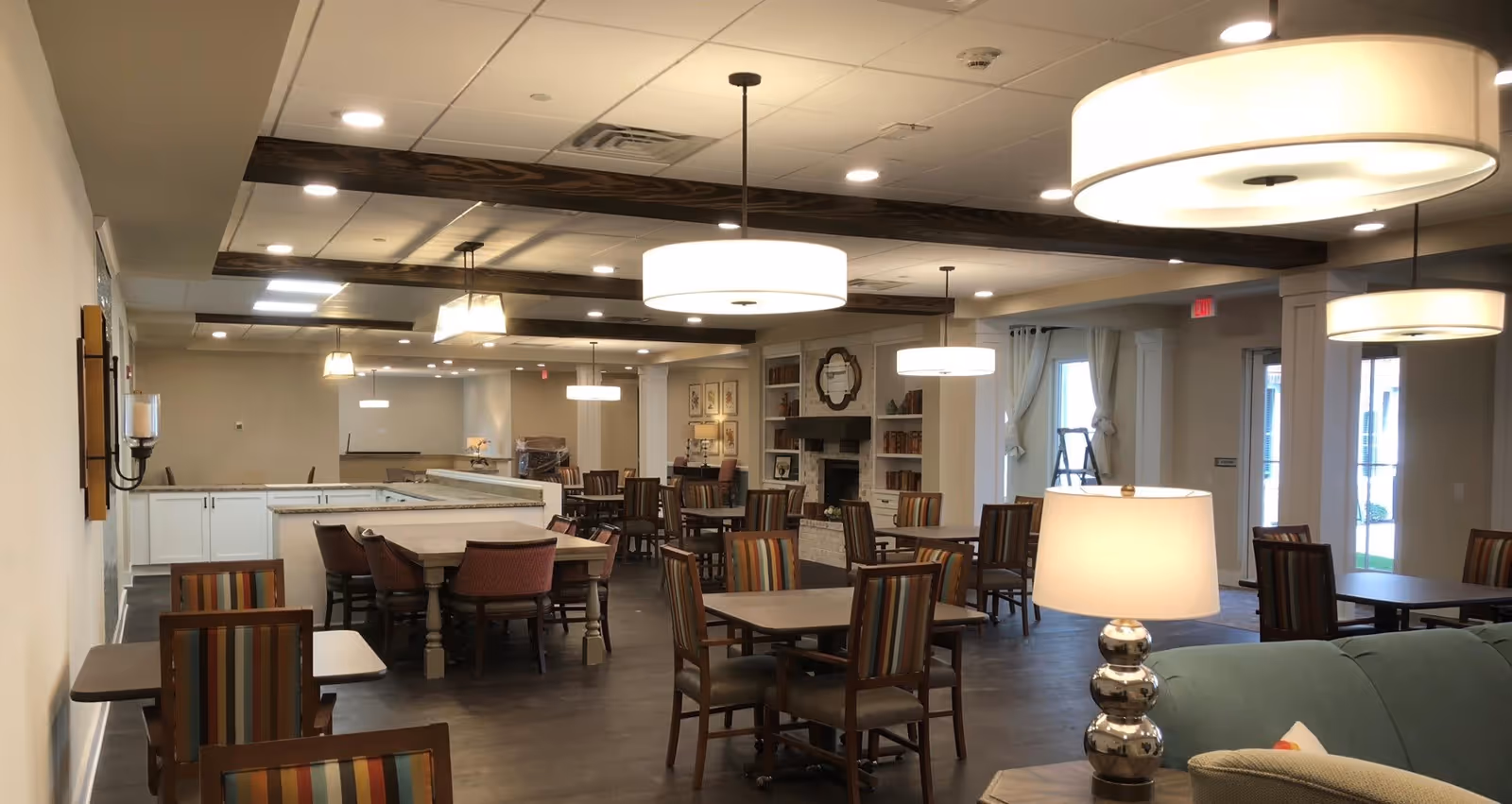 A spacious dining area in a senior living facility featuring multiple tables and chairs with striped upholstery. The room has modern pendant lighting, a fireplace with built-in shelves, and large windows with curtains allowing natural light. The ceiling has wooden beams and recessed lighting, and there is a cozy seating area with a lamp in the foreground.