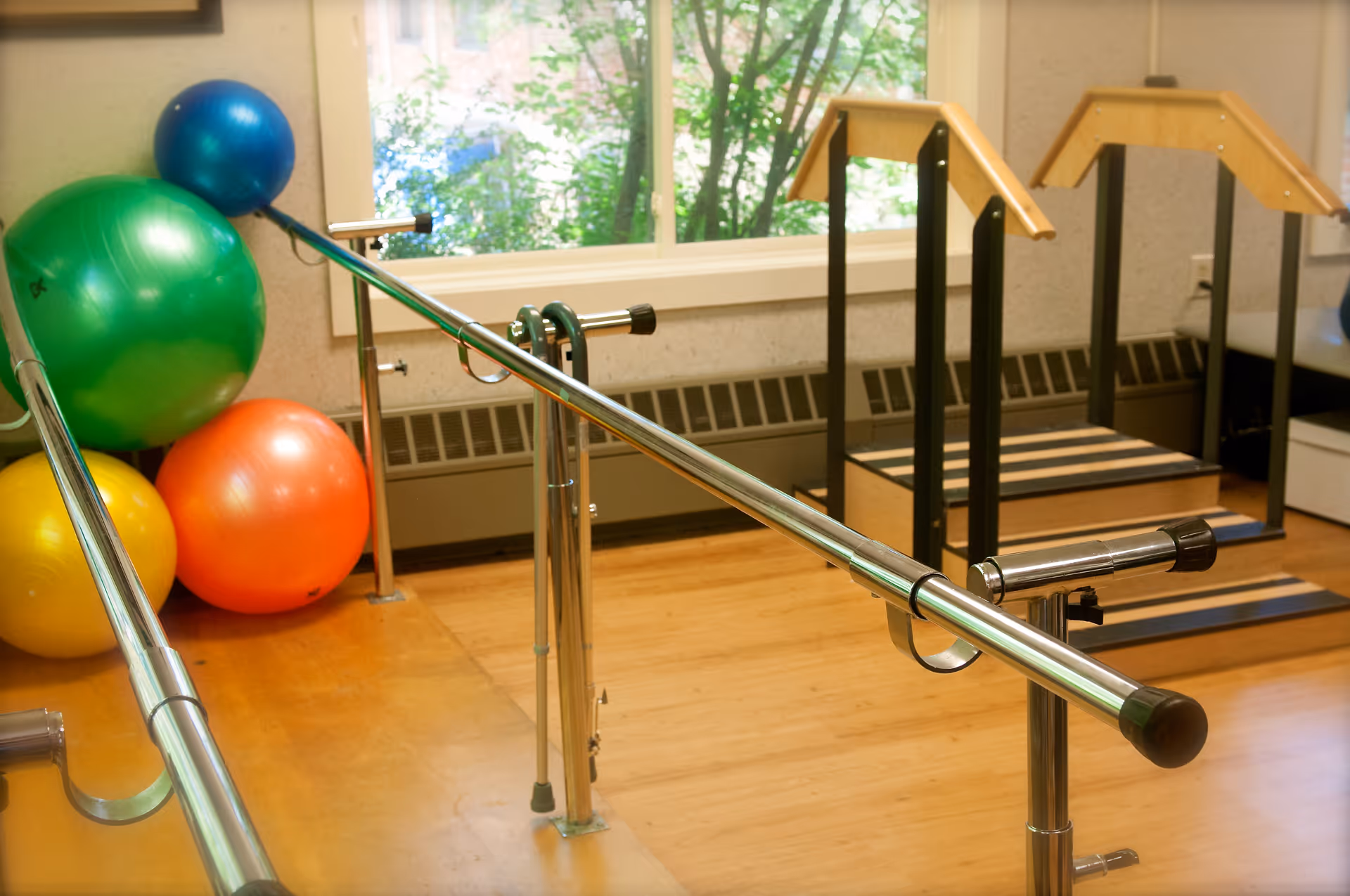 A physical therapy room with parallel bars for walking exercises, colorful exercise balls stacked in the corner, and wooden steps with handrails near a large window showing greenery outside.