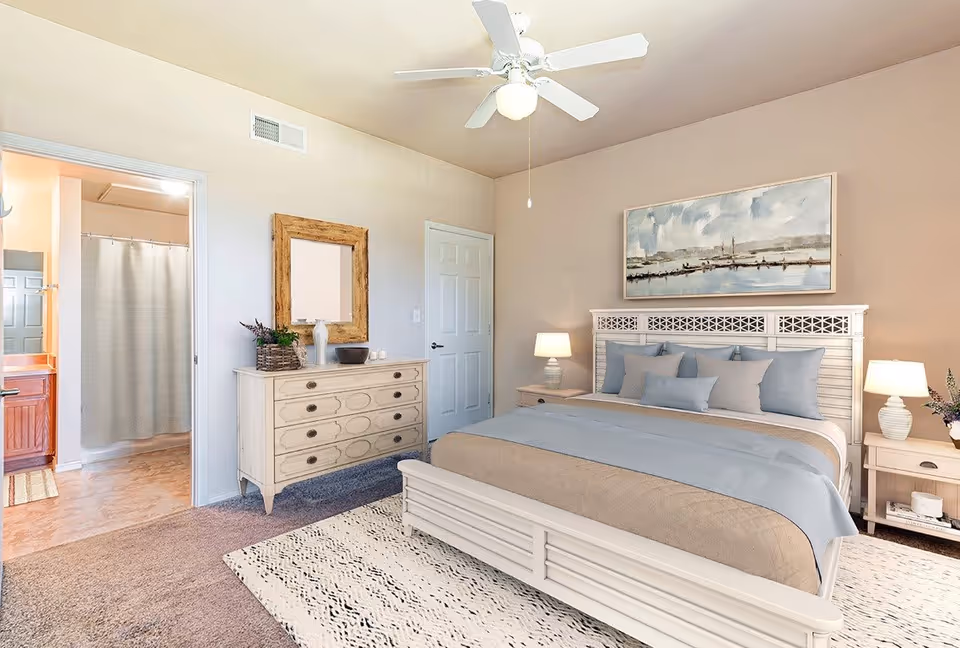 A bright and cozy bedroom featuring a large bed with beige and light blue bedding, two bedside tables with lamps, a dresser with a mirror above it, and a ceiling fan. An open door reveals a bathroom with a shower curtain and vanity.