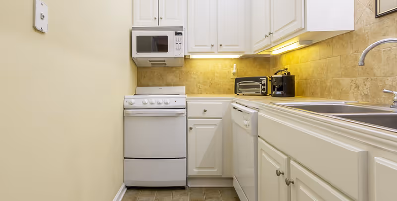 Small kitchen area with white cabinets, a white stove, microwave, dishwasher, and a double sink. There is a toaster oven and coffee maker on the countertop, with under-cabinet lighting illuminating the beige tiled backsplash.