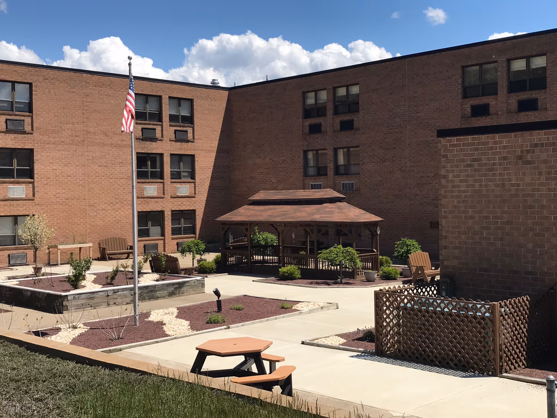 Outdoor courtyard area of a brick building with a gazebo, benches, a flagpole with an American flag, and landscaped garden beds under a partly cloudy sky.