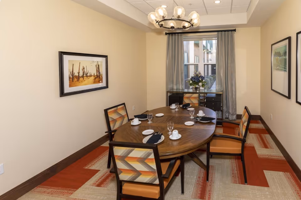 A small dining room with a wooden oval table set for four people with white cups, plates, glasses, and black napkins. Four chairs with patterned upholstery surround the table. The room has beige walls, a window with gray curtains, a chandelier with multiple glass globes, and framed artwork on the walls. A small cabinet with a flower arrangement is placed under the window.