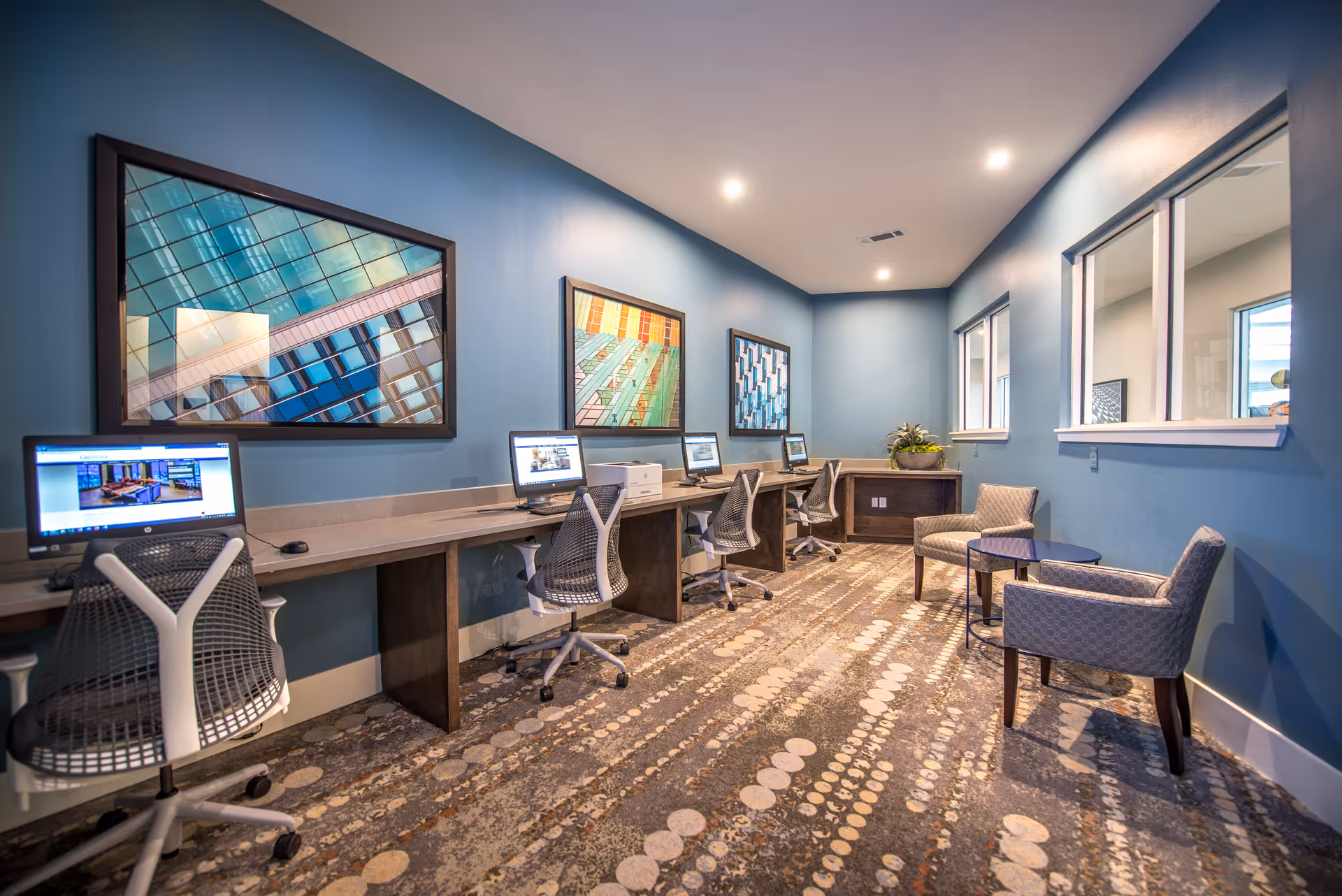 A blue-walled business center with a row of computer workstations, rolling chairs, artwork, and a small seating area.