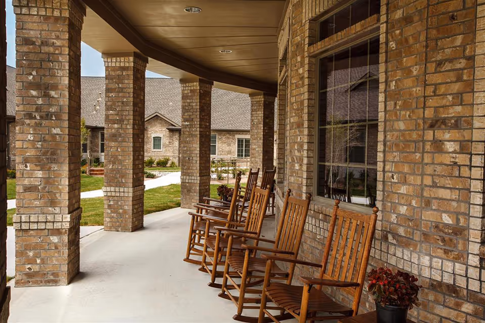 Covered outdoor porch area with several wooden rocking chairs lined up against a brick wall. The porch overlooks a landscaped garden with grass and a paved walkway. The building exterior is made of light brown bricks with large windows.