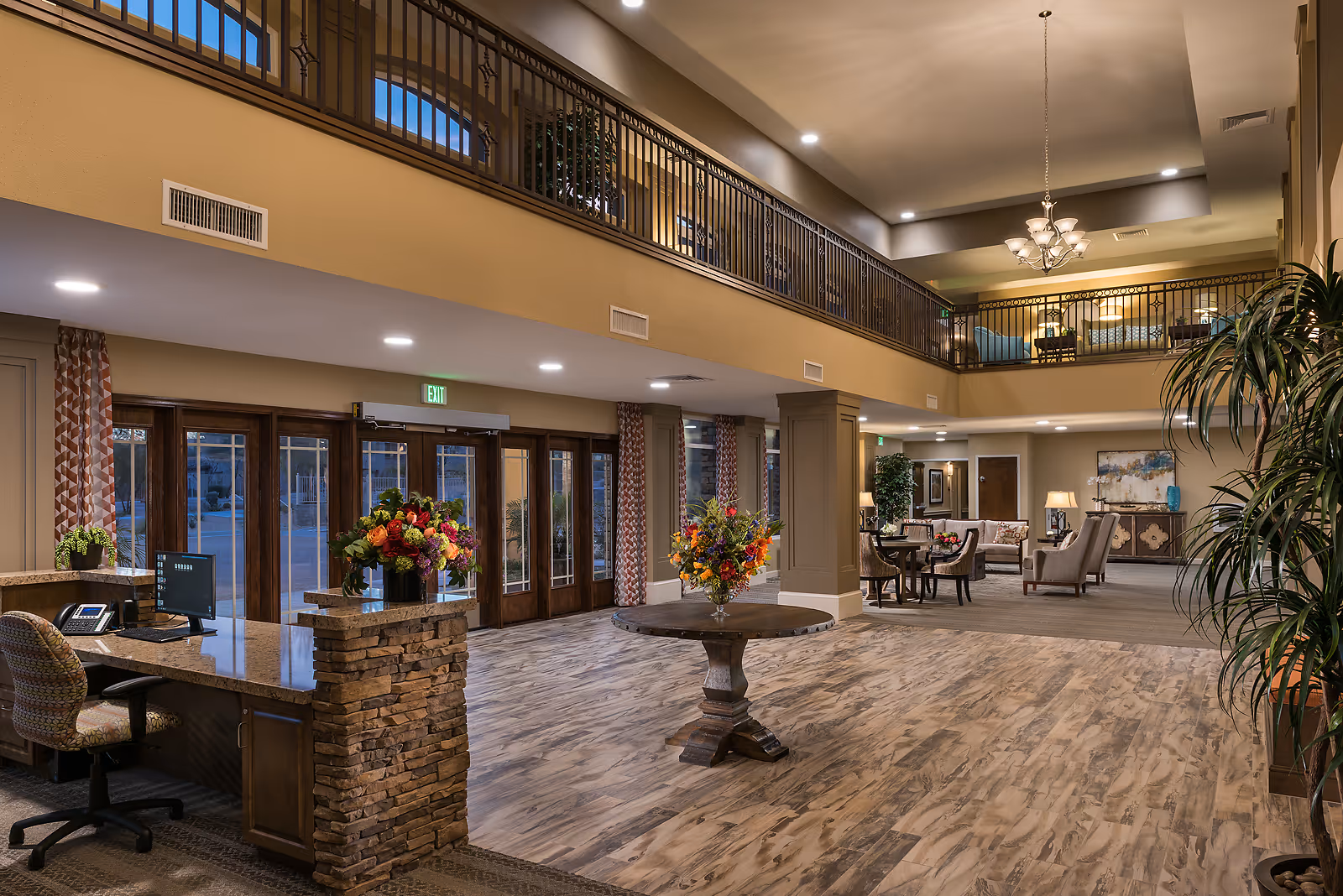 Spacious and well-lit assisted living facility lobby with a wooden reception desk on the left, a round table with a floral arrangement in the center, and seating areas with chairs and tables in the background. The lobby features high ceilings with a chandelier, large windows, and a second-floor balcony overlooking the area.