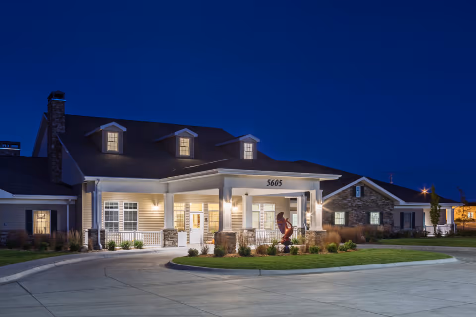 Exterior view of CountryHouse Residence senior living facility at night, showing a well-lit entrance with white pillars, stone accents, and a circular driveway with landscaping and a sculpture in front.