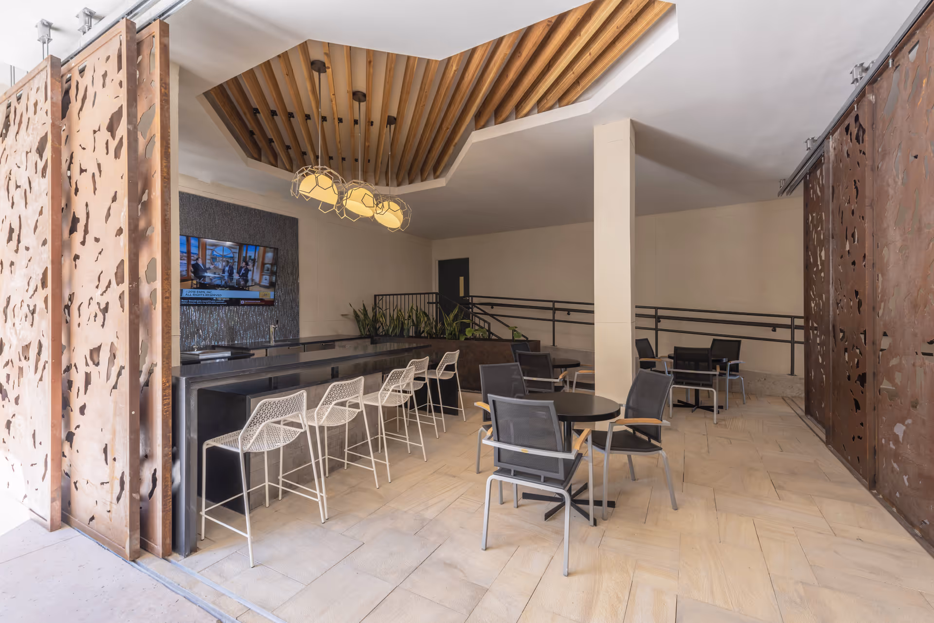 Indoor communal seating area with a long bar and white bar stools, round tables with chairs, and decorative ceiling panels.
