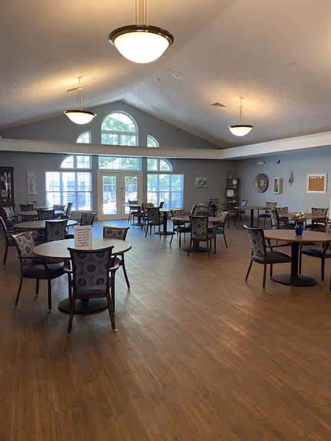A spacious dining room with multiple round tables and chairs arranged neatly on a wooden floor. Large windows with arched tops allow natural light to fill the room. The ceiling is vaulted with three hanging light fixtures. The walls are painted gray and decorated with a few framed pictures and a clock.