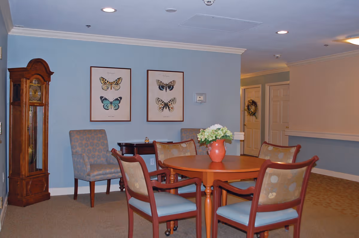 A cozy interior room with a round wooden table surrounded by four cushioned chairs. A vase with white flowers sits on the table. Behind the table, there is a wooden grandfather clock and two framed butterfly prints hanging on a light blue wall. A patterned armchair is placed near the wall, and a hallway with white doors and a wreath is visible in the background.