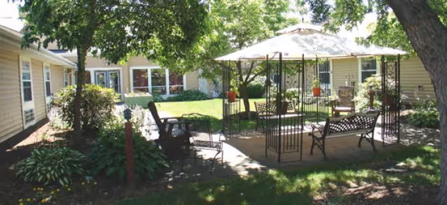 Outdoor patio area at Brookdale Painted Post with metal benches and chairs under a large umbrella, surrounded by trees and greenery, adjacent to a single-story building with multiple windows.