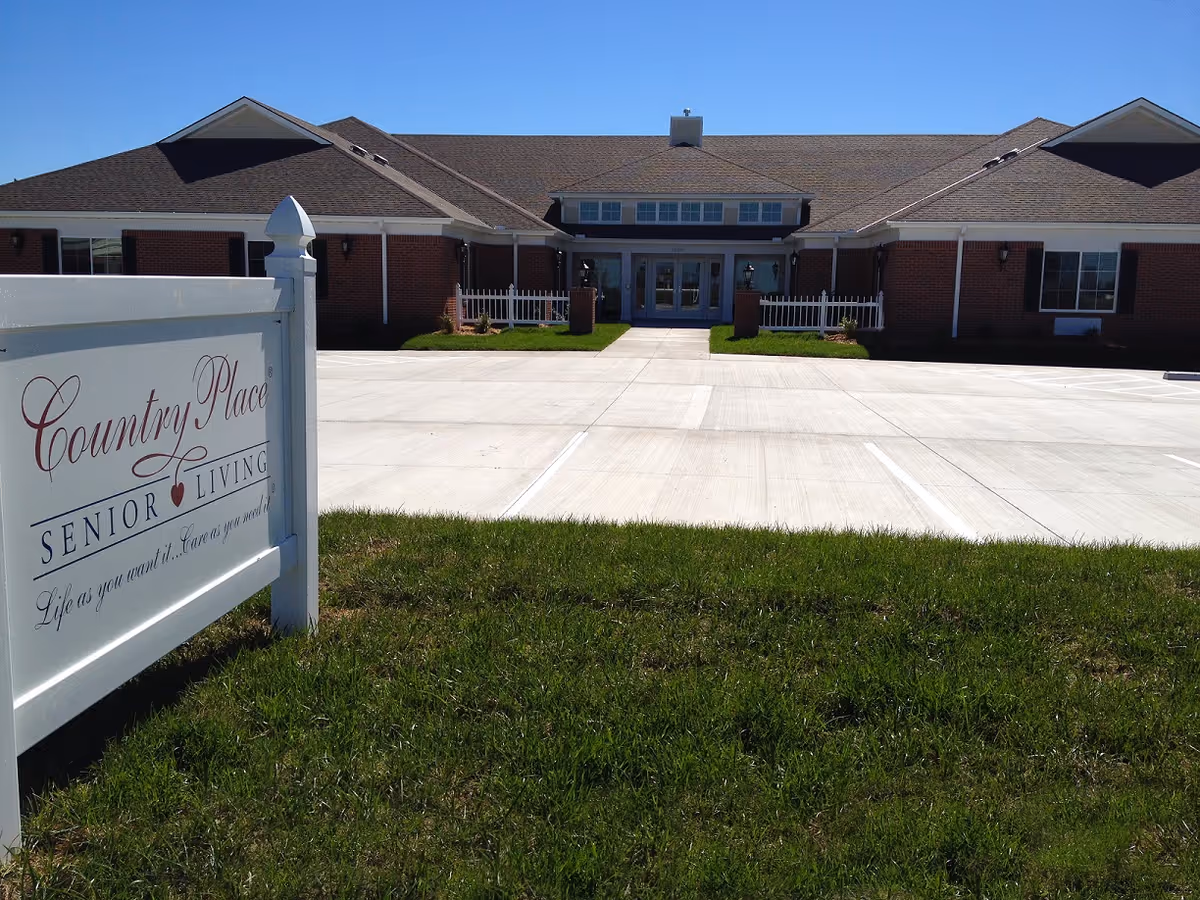 Front entrance of a single-story brick senior living building with a paved parking lot and a white sign in the foreground.