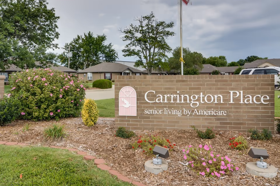 Brick entrance sign reading 'Carrington Place senior living by Americare' surrounded by landscaping and low residential buildings in the background.