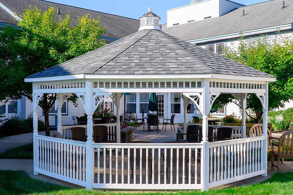 A white wooden gazebo with a shingled roof situated in a garden area with green grass and trees. Inside the gazebo, there are several chairs and tables, and in the background, a white building with multiple windows is visible.