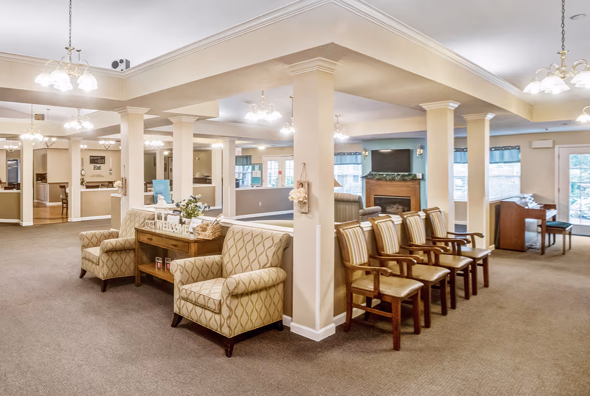 A spacious and well-lit common area in a senior living facility featuring comfortable armchairs, a row of wooden chairs with cushions, a wooden table with decorative items including a welcome sign and flowers, a fireplace with a TV mounted above it, and a piano near large windows with blue valances.