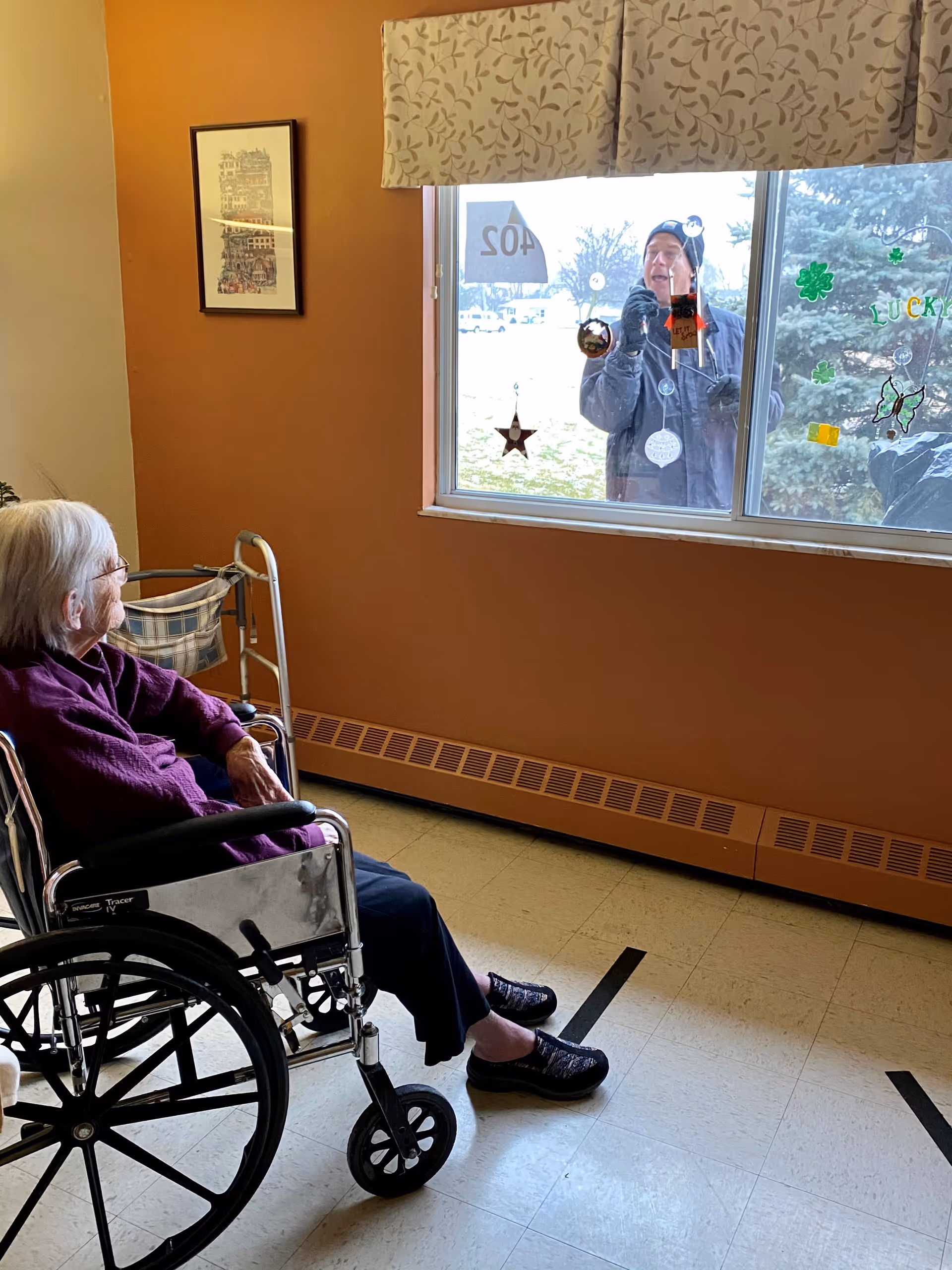 An elderly woman in a wheelchair inside a warm-toned room looks out a decorated window at a person standing outside.