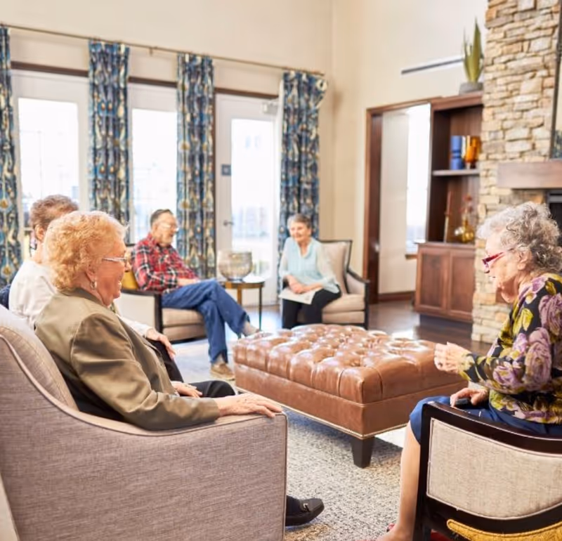 A group of elderly people sitting and conversing in a well-lit living room with large windows, patterned curtains, a stone fireplace, and a large tufted leather ottoman in the center.