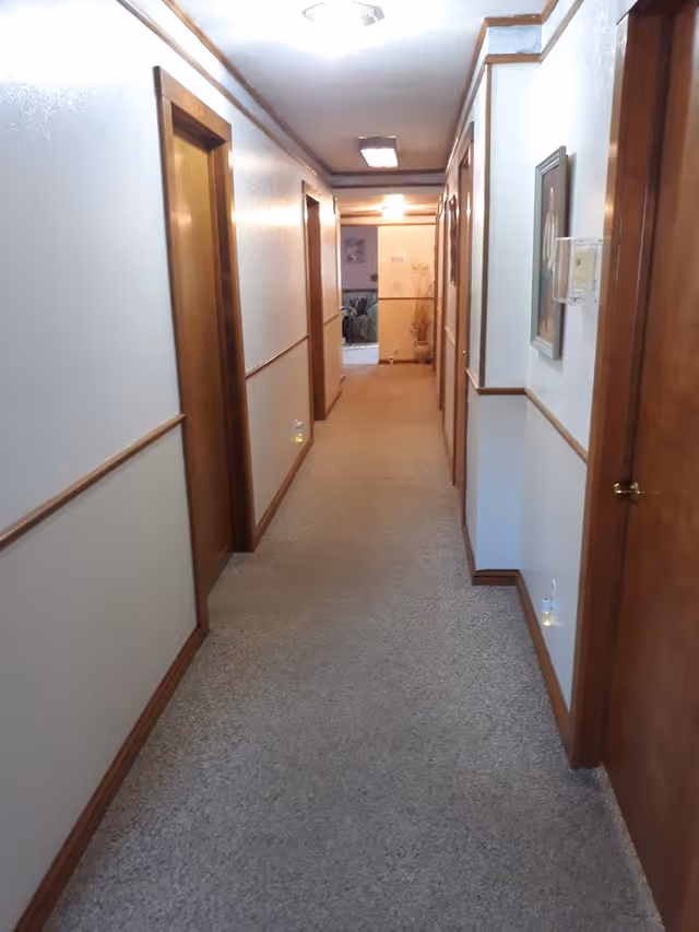 A long indoor hallway with beige carpet and white walls trimmed with wood. Several wooden doors line both sides of the hallway. The ceiling has recessed lighting and a framed picture hangs on the right wall. At the end of the hallway, a room with furniture and a plant is visible.