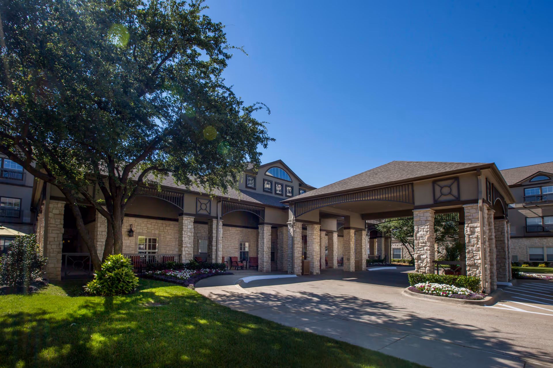 Exterior view of The Reserve at North Dallas Assisted & Senior Living facility showing a covered entrance with stone pillars, landscaped greenery, and a clear blue sky.