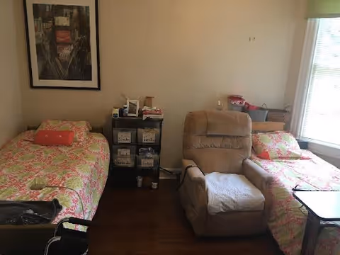 A small room with two single beds on opposite sides, each covered with matching floral bedspreads. Between the beds is a small black shelving unit holding various personal items. A beige recliner chair is positioned between the beds. A large window with blinds allows natural light into the room, and a framed picture hangs on the wall above one of the beds.