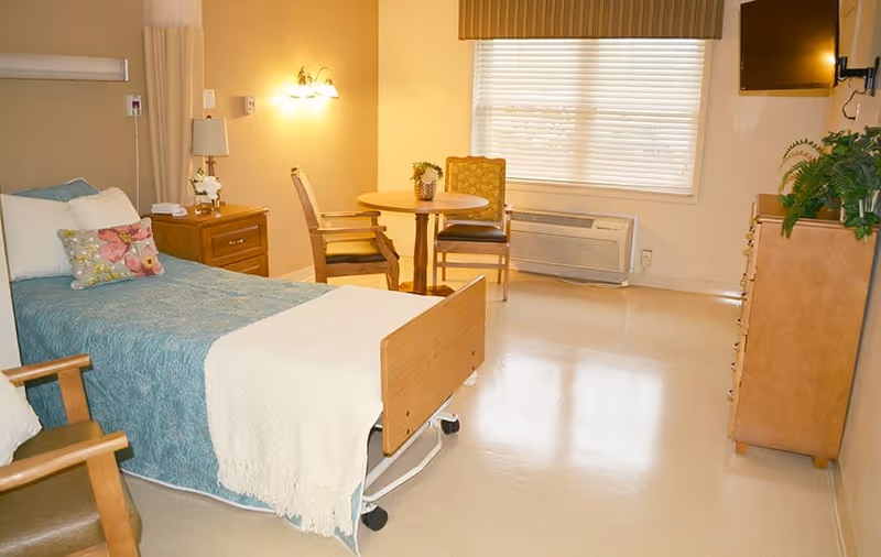 A bright nursing facility bedroom with a single bed covered in a teal blanket and white throw, a wooden nightstand with a lamp and flowers, two wooden chairs around a small round table, a window with blinds, an air conditioning unit below the window, a wooden dresser with a plant on top, and a wall-mounted TV.