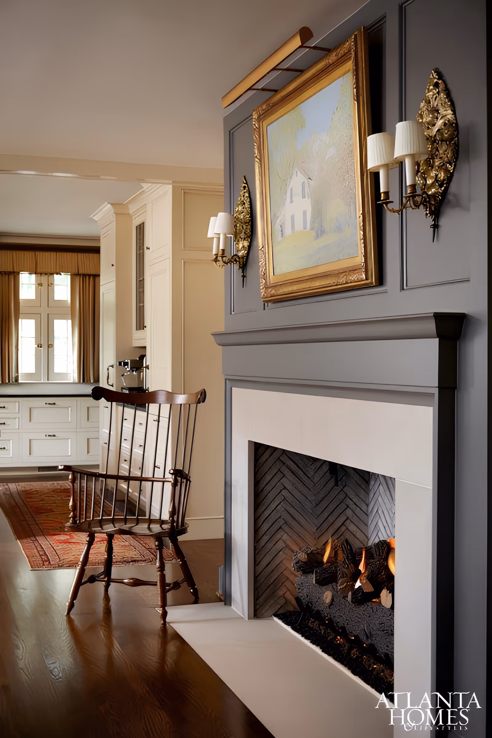 Cozy interior space featuring a lit fireplace with a gray mantel, flanked by two ornate gold wall sconces with white lampshades. Above the fireplace hangs a framed painting of a house in a natural setting. To the left, a wooden rocking chair sits on a polished hardwood floor, with a red patterned rug leading to a cream-colored kitchen area with cabinetry and a window with beige curtains.