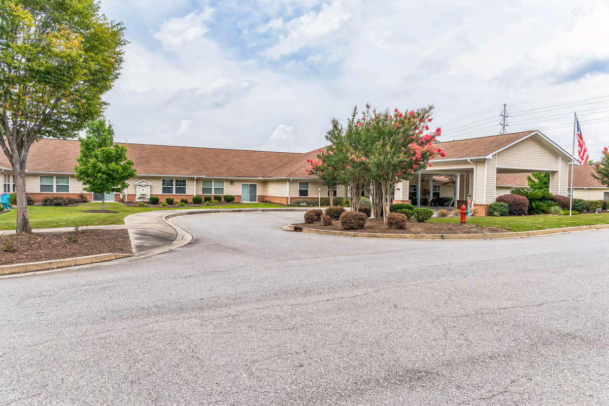 Single-story senior living facility with a covered entrance, circular driveway, landscaped islands, and an American flag.