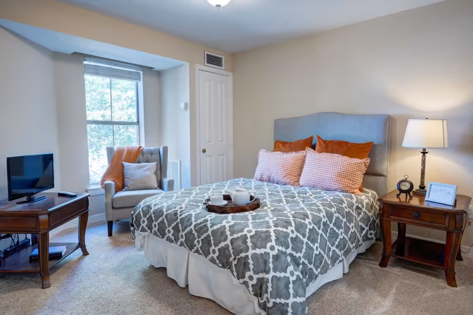 A cozy bedroom with a large bed covered in a gray and white patterned comforter and four pillows in orange and pink patterns. There is a wooden nightstand with a lamp, an alarm clock, and a framed picture on the right side of the bed. A wooden table with a small flat-screen TV and remote is on the left side near a window with a gray armchair draped with an orange throw blanket and a gray pillow. The room has beige walls and carpeted floor.