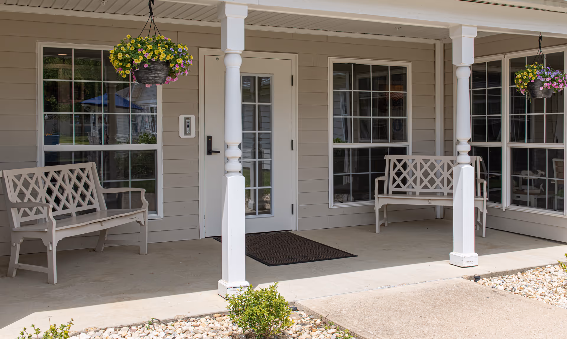 Covered porch area with two beige benches on either side of a white door with glass panels. There are two white columns supporting the porch roof and hanging baskets with yellow and purple flowers. The porch floor is concrete with a black doormat in front of the door. Windows with white frames are on either side of the door.