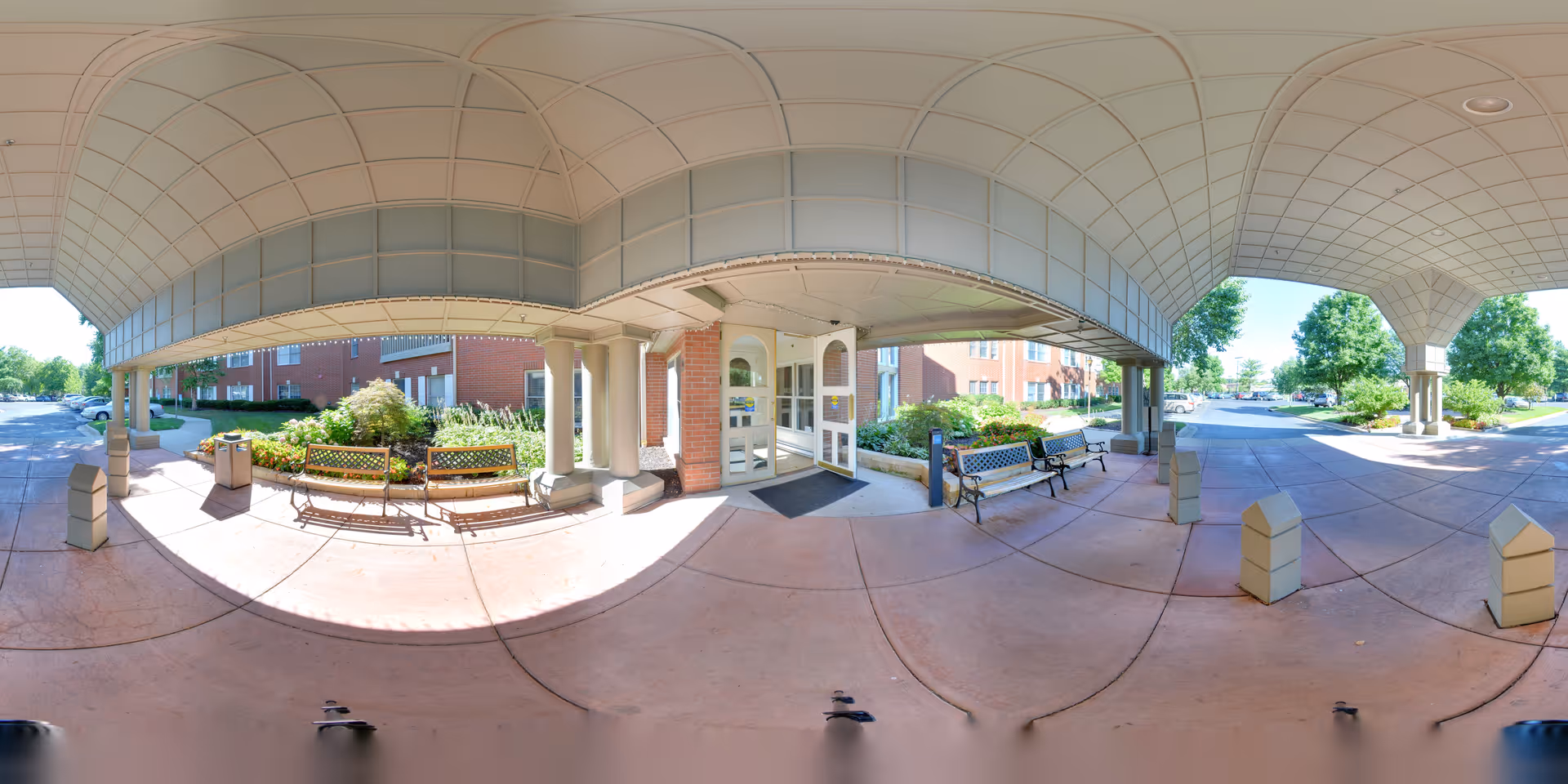 Covered entrance area of The Forum at Overland Park senior living facility with benches, columns, and a glass door entrance. The area is shaded by a large overhead canopy, with a brick building and landscaped greenery visible in the background. The driveway and parking area are also visible under clear daylight.