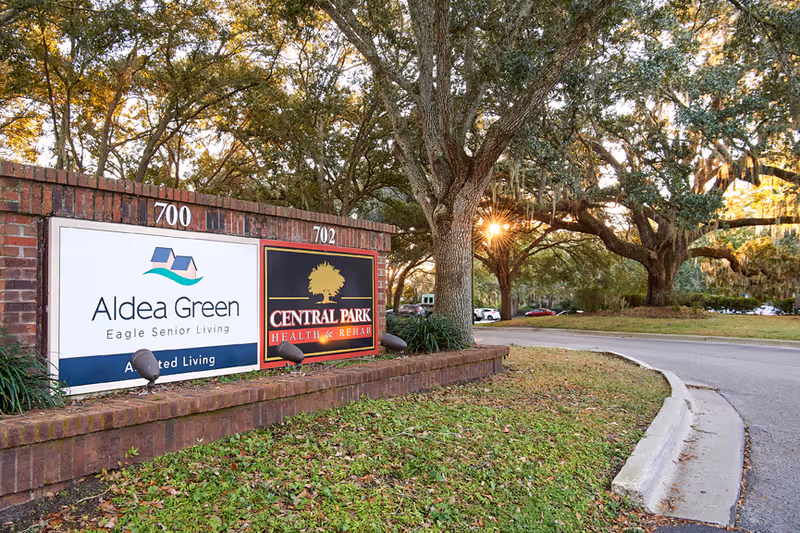 Brick entrance sign for Aldea Green and Central Park Health & Rehab next to a tree-lined driveway at sunset.
