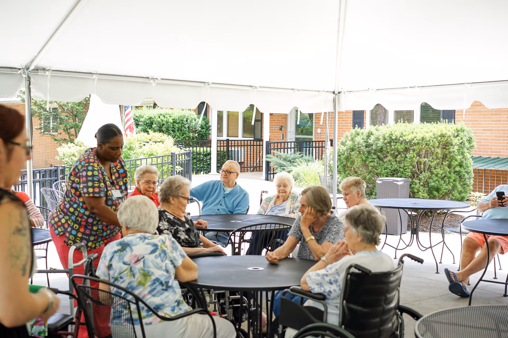 A group of elderly people sitting around outdoor tables under a white canopy tent at a nursing home. A caregiver in colorful scrubs is assisting one of the residents. The setting includes greenery and a brick building in the background.
