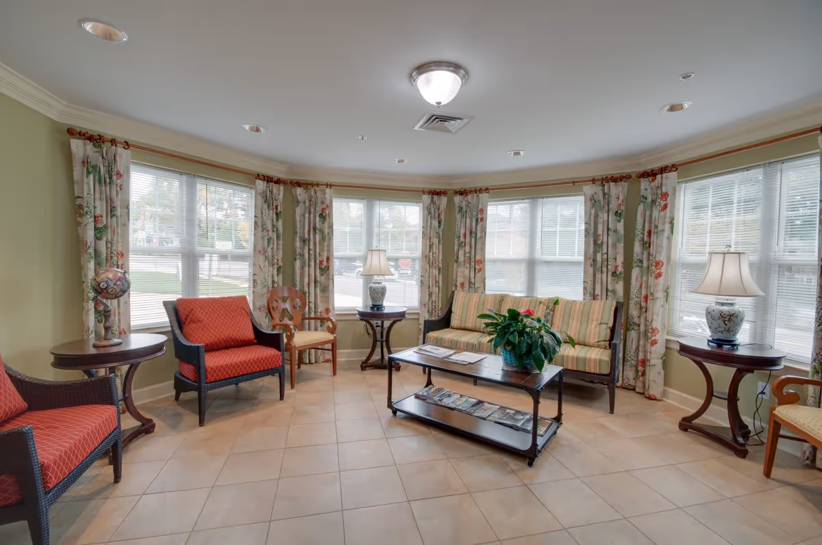 Bright communal living room with chairs, a striped sofa, a coffee table, lamps, and large windows with floral curtains.