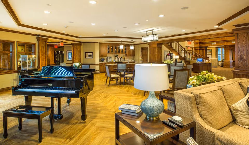 A spacious and well-lit common area in a senior living facility featuring a black grand piano with a matching bench, a beige sofa with cushions, a wooden side table with a blue ceramic lamp, books, and remote controls. In the background, there are several wooden dining tables with chairs, a kitchenette area with cabinets and coffee machines, and wooden columns and trim throughout the room.