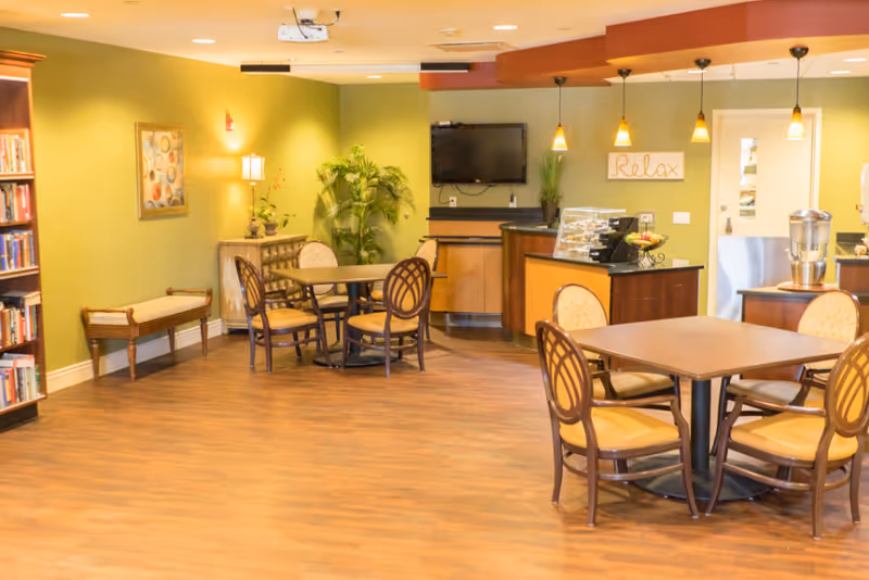 A cozy senior living common area with wooden flooring, several round tables with cushioned chairs, a bookshelf filled with books, a bench, a small cabinet with a lamp and plant, a wall-mounted TV, and a counter with a display case and a water dispenser. The walls are painted green and decorated with a framed picture and a sign that says 'Relax'.