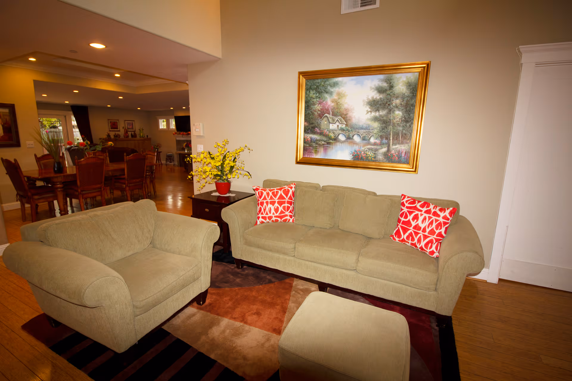 A cozy living room area with a beige sofa and matching armchair, both adorned with red and white patterned throw pillows. A small ottoman is in front of the sofa. There is a wooden side table with a red vase holding yellow flowers. On the wall above the sofa hangs a framed painting of a serene landscape featuring a stone bridge over a river surrounded by trees and flowers. In the background, a dining area with a wooden table and chairs is visible, along with warm lighting and hardwood floors.