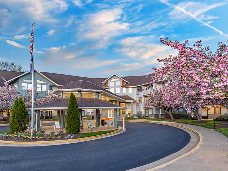 Exterior view of the Virginian senior living facility with a curved driveway, a covered entrance, blooming pink cherry blossom trees, and an American flag on a flagpole under a partly cloudy sky.