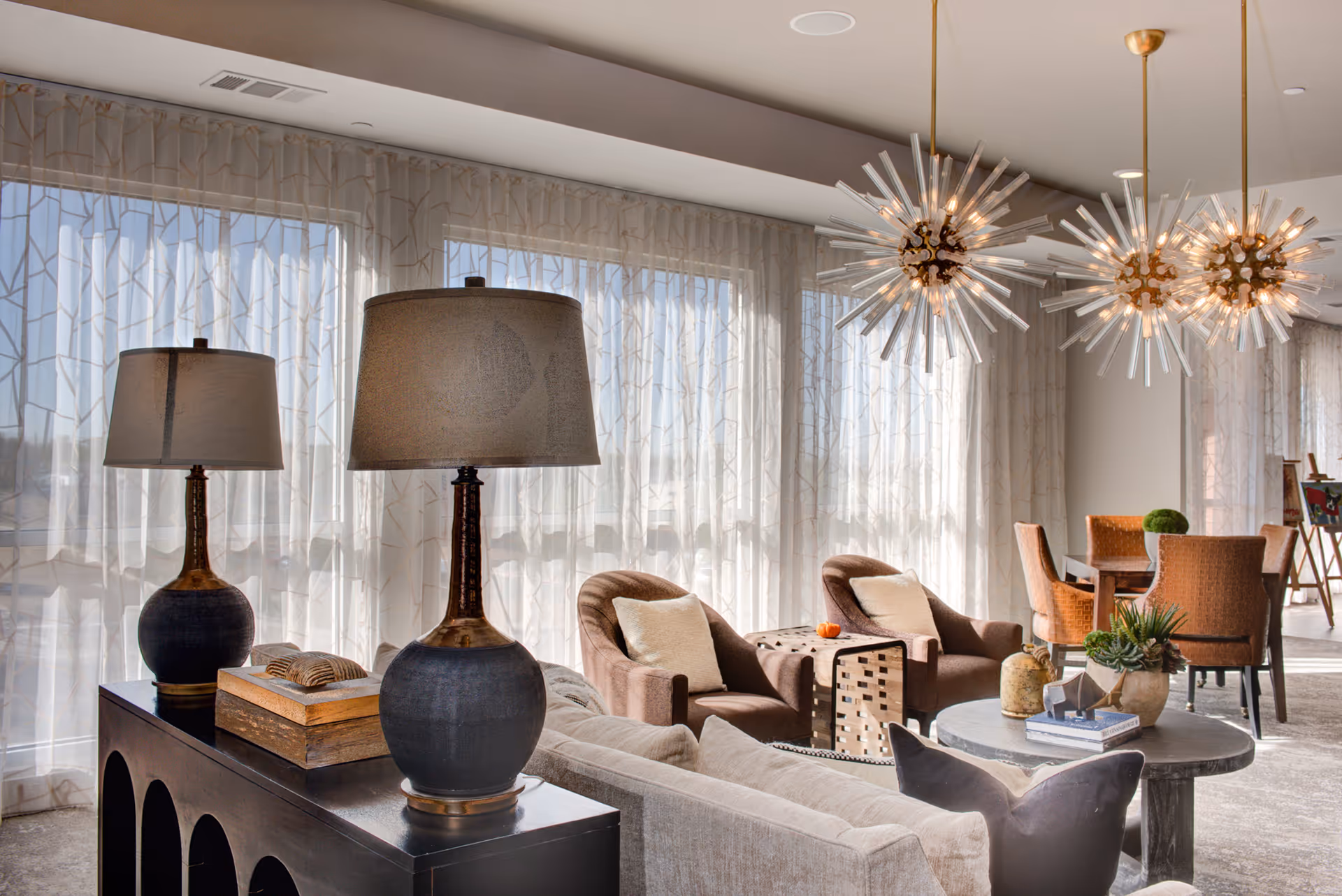 A bright and modern living room area with large windows covered by sheer curtains. The room features a beige sofa with pillows, two brown armchairs with light cushions, a round coffee table with decorative items, and a black console table with two matching lamps. In the background, there is a dining table with orange upholstered chairs and three unique starburst pendant lights hanging from the ceiling.