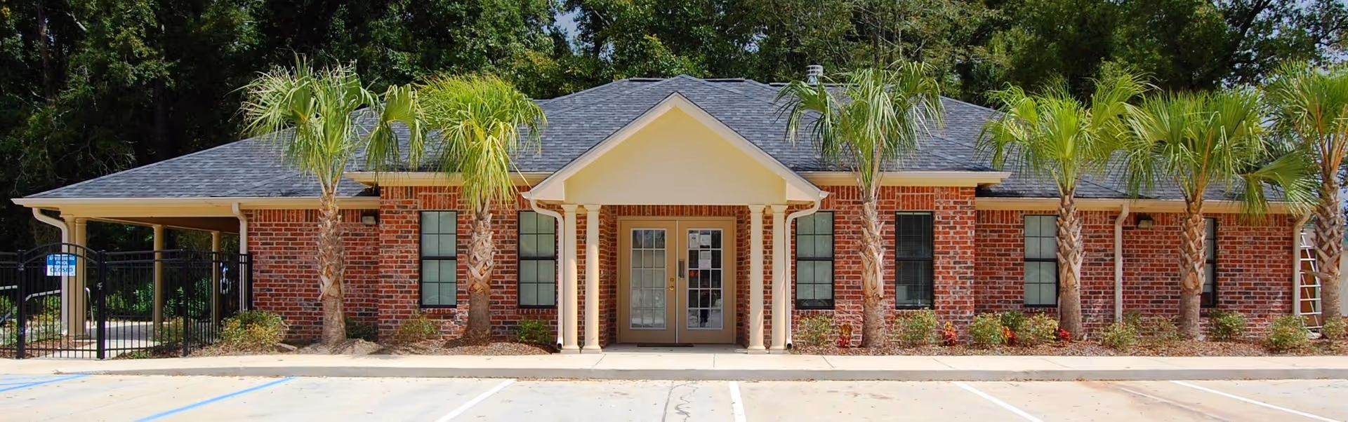 Front exterior view of a single-story brick building with a covered entrance supported by columns, surrounded by palm trees and landscaping, with a parking area in front.