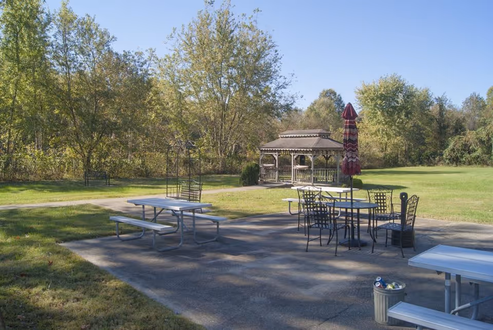 Outdoor patio with picnic tables, metal dining chairs and a closed umbrella near a gazebo on a grassy lawn bordered by trees.
