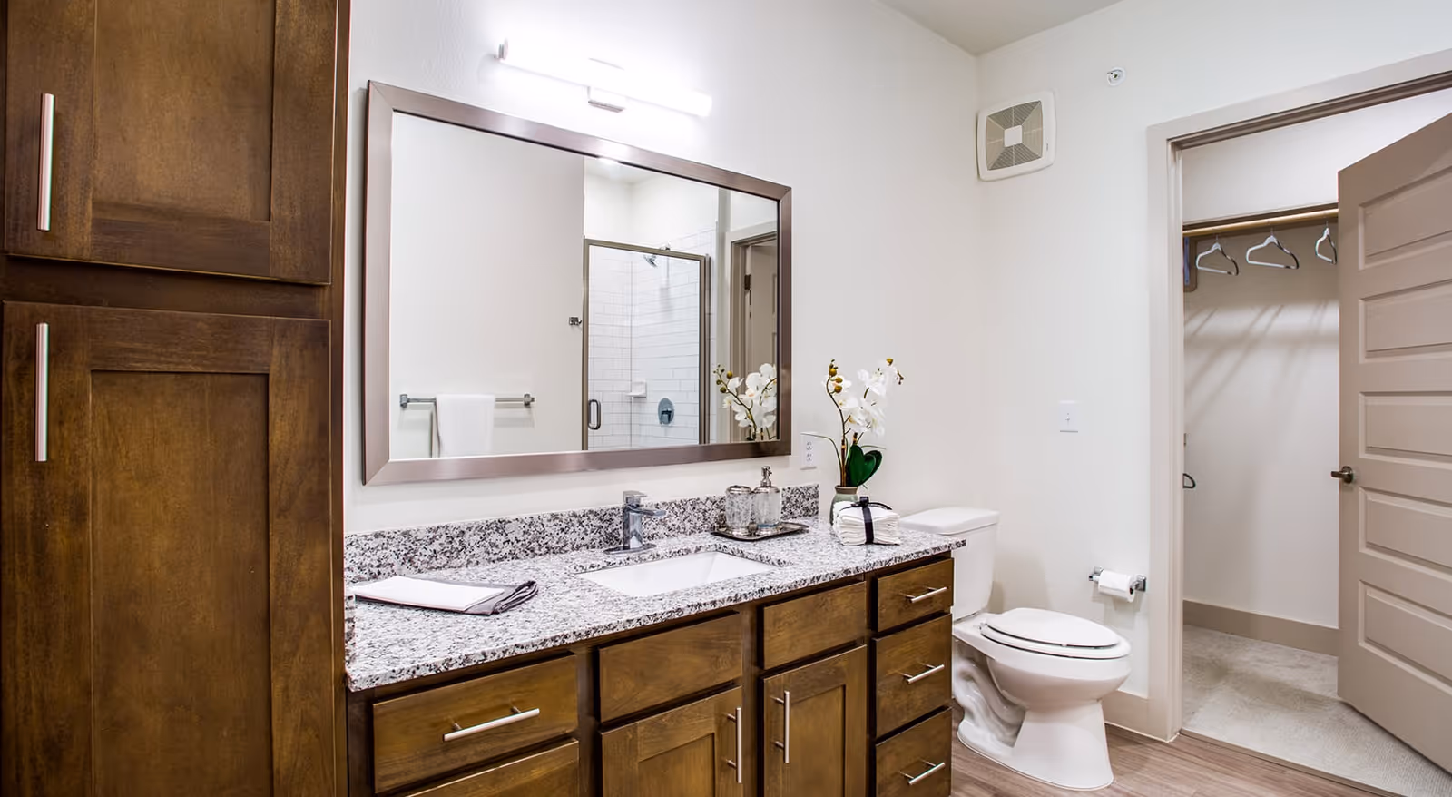 A clean and modern bathroom featuring a granite countertop with a sink, wooden cabinets, a large rectangular mirror, a toilet, and an open door leading to a walk-in closet with hangers. The bathroom has white walls and a glass-enclosed shower visible in the mirror.