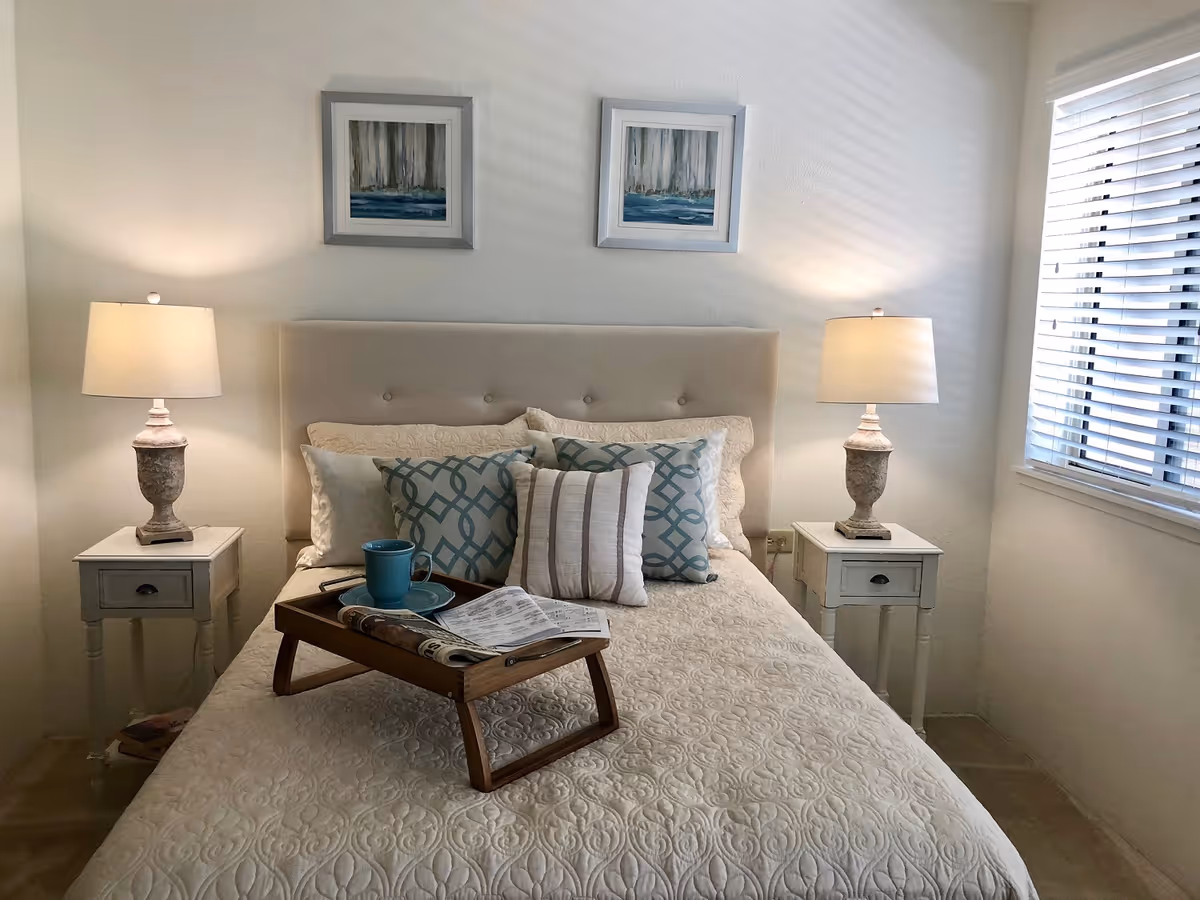 A neatly made bed with a beige quilted bedspread and multiple decorative pillows in shades of beige and blue. There is a wooden tray on the bed holding a blue cup and saucer, a newspaper, and a pen. On either side of the bed are white nightstands with matching table lamps. Two framed abstract paintings hang above the bed. A window with white blinds is on the right side of the room.