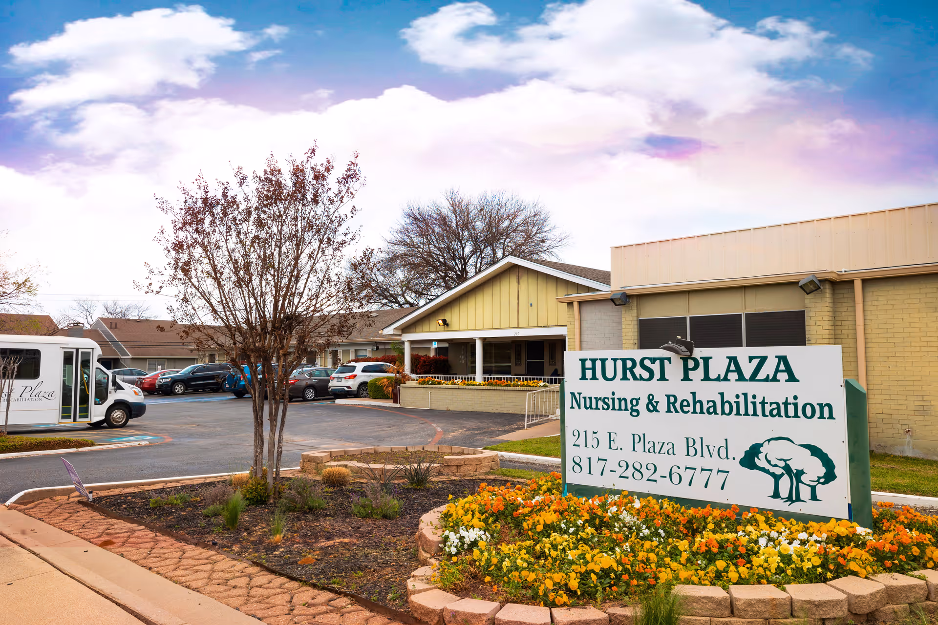 Front exterior of Hurst Plaza Nursing & Rehabilitation showing the entrance sign, flowerbed, driveway and parked vehicles.