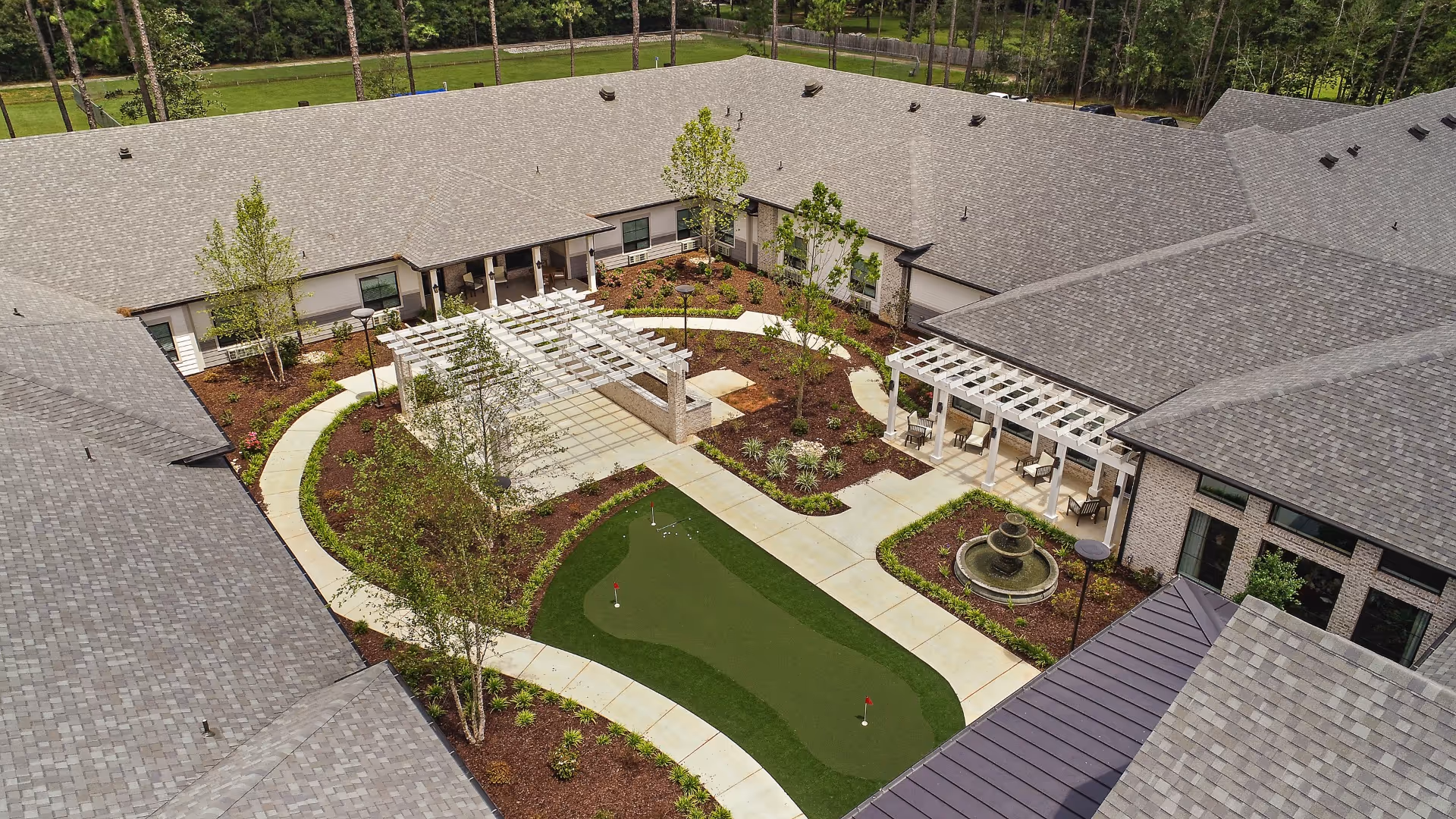 Aerial view of a central courtyard with a putting green, walkways, pergolas, a fountain and surrounding building roofs.